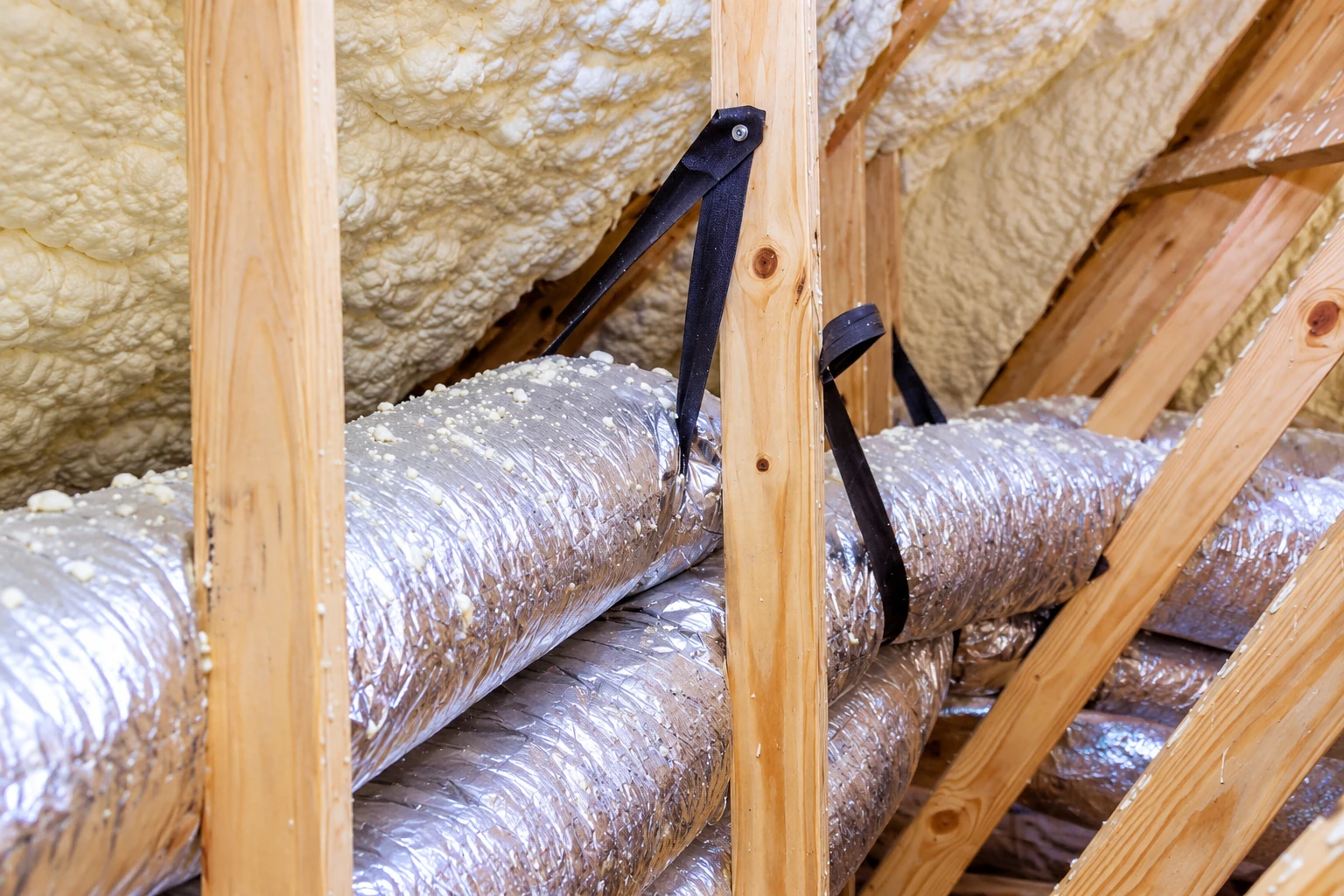 Silver flexible ducts run between wooden attic rafters beneath a roof coated with pale spray foam.