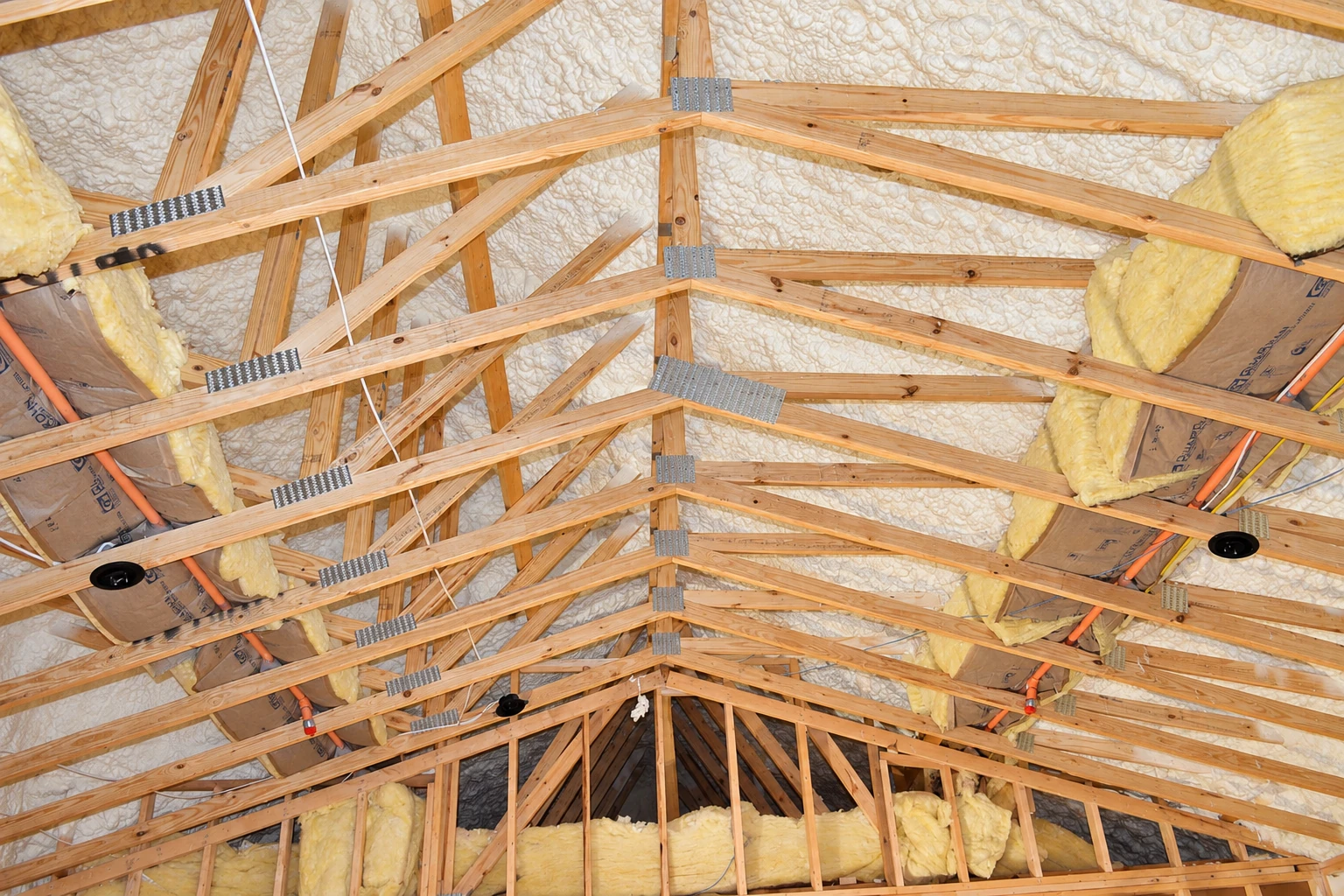 Wooden attic trusses frame pale spray foam on the roof deck, with yellow batt insulation tucked between sections.