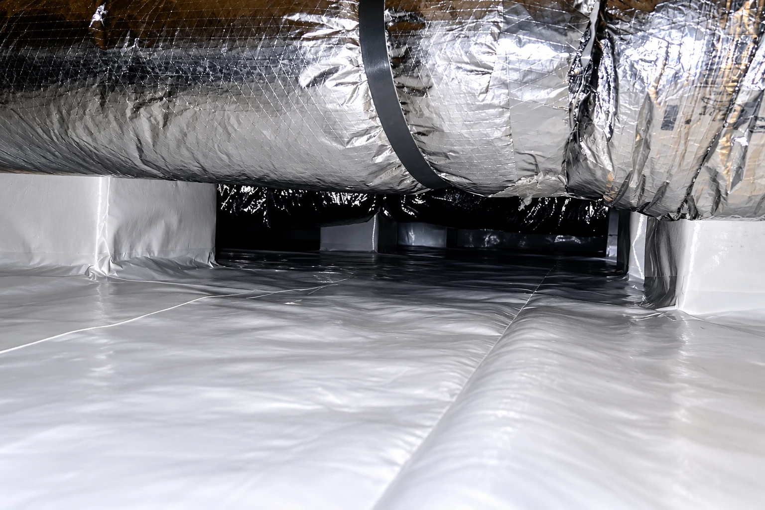 A crawl space has a shiny white vapor barrier across the floor and silver-wrapped ductwork overhead.